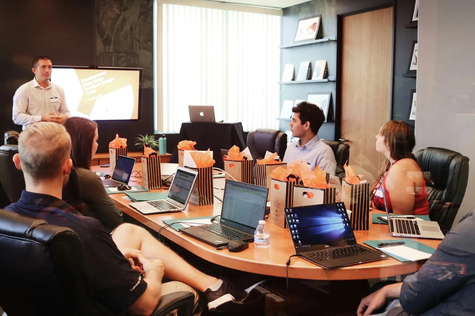 Team sitzt mit Laptops an einem Besprechungstisch, vorne präsentiert eine Person