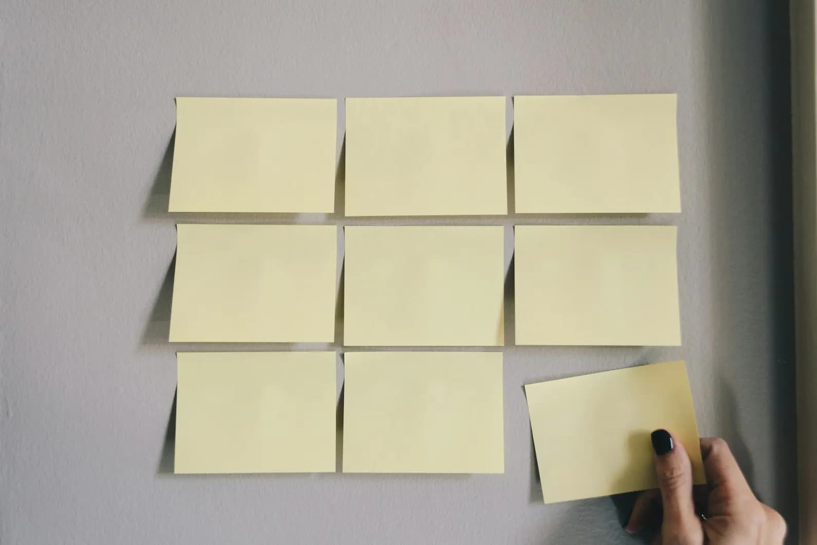 Neun leere gelbe Haftnotizen an einer grauen Wand, eine Hand klebt die letzte auf
